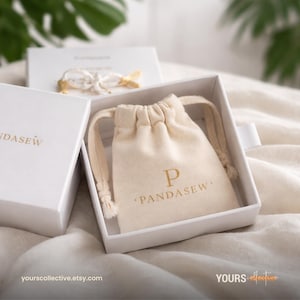 May include: A white box with the word "PANDASEW" in gold lettering. Inside the box is a small beige drawstring bag with the letter "P" and the word "PANDASEW" printed in gold. The background includes a white surface and a box with a bow.