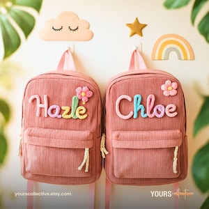 May include: Two pink corduroy backpacks with personalized names in colorful yarn. Each backpack has a front pocket, a flower applique, and a top handle. Decorative cloud, star, and rainbow wall hangings are above the backpacks.