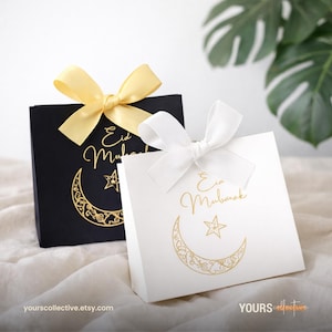 Peut inclure: Deux boîtes cadeaux, une noire et une blanche, avec des motifs dorés et des croissants de lune. Chaque boîte a un nœud en satin, un jaune et un blanc. Le texte "Eid Mubarak" est visible sur les deux boîtes.