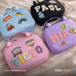 May include: Four colorful, rectangular bags with handles. Each bag features a name in large, embroidered letters and decorative patches. The bags are in light blue, black, lavender, and pink. The bags have white and light blue striped handles.