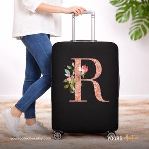 Puede incluir: Funda de maleta negra con una gran letra "R" con purpurina oro rosa y un diseño floral de rosas rosas y hojas verdes. La funda está sobre una maleta con ruedas.
