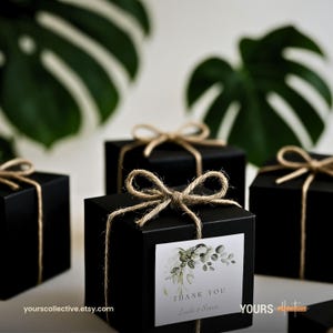 May include: Several black gift boxes tied with twine bows. Each box has a small white label with the words "THANK YOU" and a botanical design. The boxes are arranged on a white surface with large green leaves in the background.