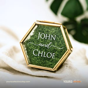 Peut inclure: Boîte à bagues hexagonale en verre avec bordure dorée, remplie de mousse verte. Les noms "JOHN and CHLOE" sont affichés en écriture blanche. Un article décoratif pour les mariages ou les occasions spéciales.