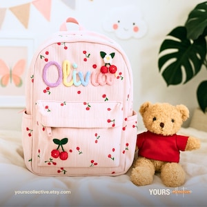 May include: A pink corduroy backpack adorned with a cherry pattern and the name "Olivia" in colourful yarn letters. The backpack features a front pocket, side pockets, and a top handle. A teddy bear in a red shirt is placed beside it.