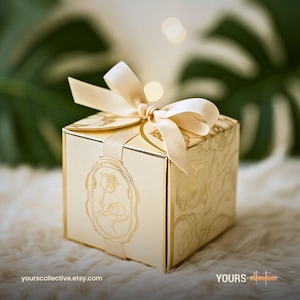 May include: A gold gift box with a cream ribbon tied in a bow. The box features a floral pattern and a decorative tag with a rose design. The box is on a white surface.
