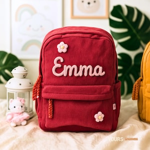 May include: A red corduroy backpack with the name "Emma" embroidered in pink script. The backpack features a front pocket, two small flower appliqués, and a braided zipper pull. A small stuffed animal and decorative lantern are in the background.