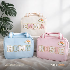May include: Three personalized lunch bags in pastel colors: cream, light blue, and pink. Each bag features a name in a varsity-style font with gold trim and a rainbow patch. The bags have matching striped handles and zippers.