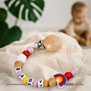 Peut inclure: Un attache-tétine pour bébé en bois avec un arc-en-ciel et le mot "BABY" épelé avec des perles de lettres. Le clip a un disque en bois et un fermoir en métal. Le clip est sur une surface blanche.