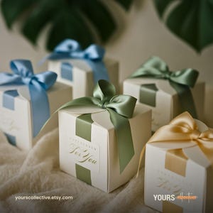 Puede incluir: Una colección de cajas de regalo cuadradas blancas, cada una adornada con un lazo de cinta de satén en tonos azul, verde salvia o dorado. Cada caja presenta una etiqueta cuadrada con el texto en relieve "For You" en letra cursiva.