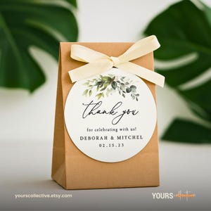 May include: A small brown paper gift bag tied with a cream-colored ribbon. A white circular tag is attached, featuring the words "Thank you" in elegant script, with botanical illustrations and the names Deborah & Mitchel, and the date 02.15.23.