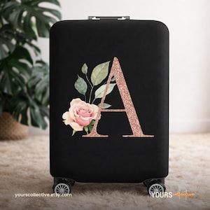 Könnte beinhalten: Schwarze Kofferhülle mit einem großen roségoldenen Glitzerbuchstaben "A" und einem Aquarell-Blumenmuster. Das Design umfasst eine rosa Rose und grüne Blätter. Die Hülle befindet sich auf einem Rollkoffer.