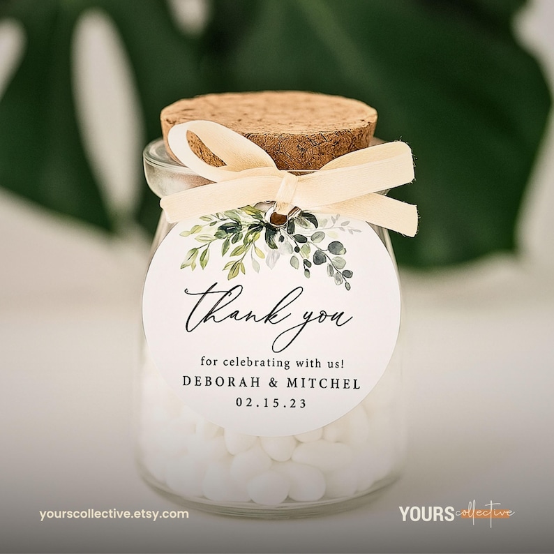 May include: A small glass jar filled with white candies, topped with a cork and tied with a cream-colored ribbon. A round tag reads "Thank you" with the names Deborah & Mitchel and the date 02.15.23, with a floral design.