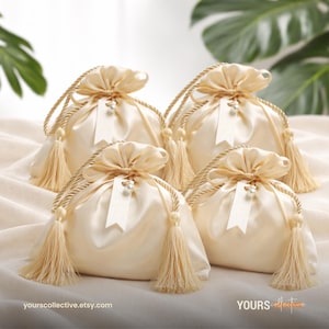 May include: Four cream satin drawstring bags with tassels and rope handles. Each bag has a small tag and a pearl accent. The bags are arranged on a cream surface. The text "YOURS collective.etsy.com" is visible.