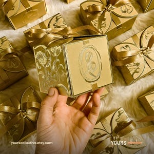 Puede incluir: Cajas de regalo doradas con diseños florales y cintas a juego. Una caja se sostiene en la mano, mostrando el diseño de tulipán en relieve. Las cajas están dispuestas sobre una superficie suave y blanca. La URL de la tienda Etsy es visible.