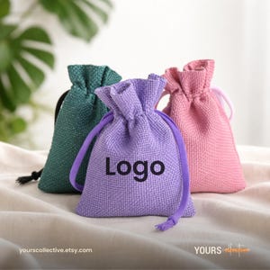 May include: Three small drawstring bags in green, purple, and pink. The purple bag has the word "Logo" printed on it. The bags are made of a textured fabric and are displayed on a neutral surface.