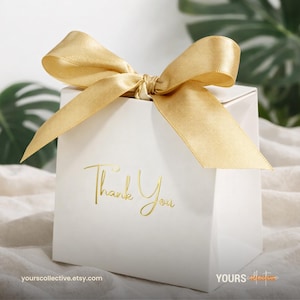 May include: A white gift box with a gold satin ribbon tied in a bow. The words "Thank You" are printed in gold script on the front of the box. The box is sitting on a white surface.
