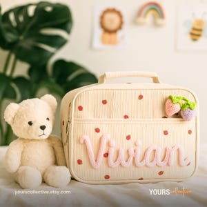 Op de afbeelding: Crèmekleurige lunchtas met een handvat, versierd met rode aardbeienaccenten en de naam "Vivian" in roze garen. Voorzien van een voorvak met twee gehaakte aardbeienbedels. Een knuffelbeer staat naast de tas.