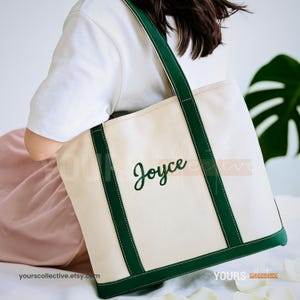 May include: Cream-colored tote bag with green trim and handles. The word "Joyce" is embroidered in green script on the front. The bag is made of canvas material and has a rectangular shape. The Etsy shop name "yourscollective.etsy.com" is printed on the bottom left.