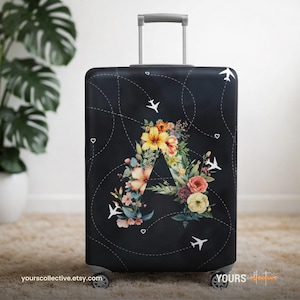 Könnte beinhalten: Schwarze Kofferhülle mit floralem "A"-Design mit gelben, rosa und orangefarbenen Blumen. Weiße Flugzeuge und gepunktete Linien zieren den schwarzen Hintergrund. Der Koffer hat einen ausziehbaren Griff und Rollen. Die Website yours collective.etsy.com ist unten aufgedruckt.