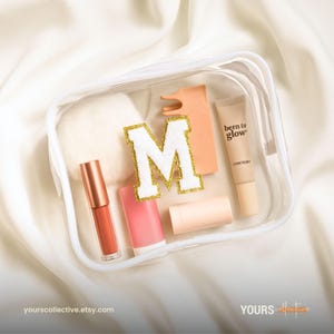Può includere: Un beauty case trasparente pieno di prodotti di bellezza. All'interno ci sono un lucidalabbra, un rossetto, un correttore, una lettera "M" color pesca con bordi glitterati dorati e altri articoli per il trucco. La borsa è su una superficie di raso bianco. Il testo "yours collective.etsy.com" è visibile.