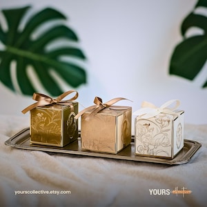 May include: Three decorative gift boxes with floral patterns and satin ribbon bows. The boxes are gold, champagne, and ivory, and are displayed on a silver tray. The Etsy shop URL is visible.