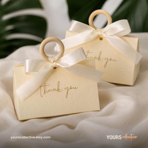 May include: Two cream-coloured gift boxes featuring a subtle embossed floral pattern and the text thank you in gold script. Each box is topped with a smooth wooden ring handle and a tied cream satin ribbon bow, creating a decorative presentation.