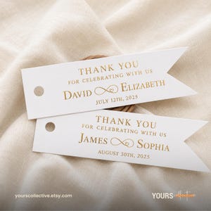Könnte beinhalten: Zwei weiße, rechteckige Papieranhänger mit einem spitz zulaufenden Ende in Flaggenform und einem runden Loch. Die Anhänger tragen goldfarbenen Text mit der Aufschrift THANK YOU FOR CELEBRATING WITH US, gefolgt von den Namen und Daten DAVID UND ELIZABETH 12. JULI 2025 sowie JAMES UND SOPHIA 30. AUGUST 2025.