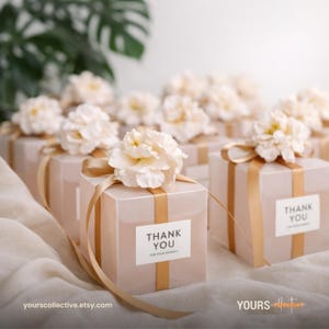 Puede incluir: Varias cajas de regalo pequeñas y transparentes con cintas doradas y flores blancas. Cada caja tiene una etiqueta que dice "THANK YOU FOR YOUR KINDNESS". Las cajas están dispuestas sobre una superficie blanca texturizada.