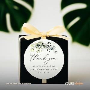 May include: A black gift box with a cream-colored ribbon and a circular label. The label features the words "Thank you" in elegant script, along with the names Deborah & Mitchel and the date 02.18.23. The label is decorated with green and gray botanical illustrations.