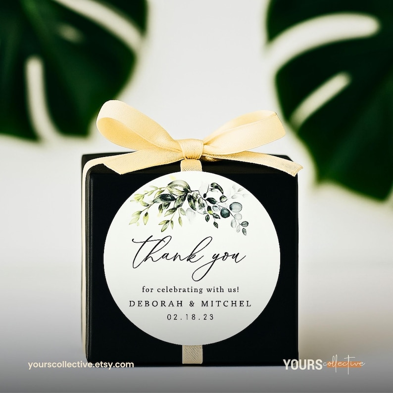 May include: A black gift box with a cream-colored ribbon and a circular label. The label features the words "Thank you" in elegant script, along with the names Deborah & Mitchel and the date 02.18.23. The label is decorated with green and gray botanical illustrations.