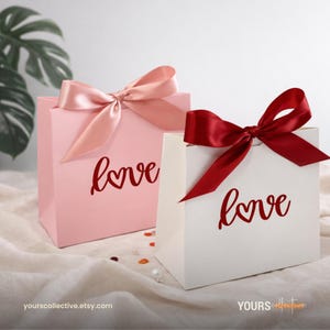 May include: Two gift bags are shown, one pink and one white. Each bag features a satin bow and the word "love". The pink bag has a pale pink bow, the white bag has a deep red bow. Ideal for gifts.