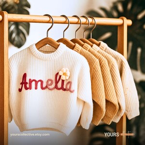 Könnte beinhalten: Eine Reihe von Kinderpullovern in Creme, Beige und Senfgelb hängen an einer Holzstange. Der erste cremefarbene Pullover ist mit dem Namen "Amelia" in Rot bestickt und hat eine Blumenapplikation.