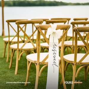 Op de afbeelding: Houten stoelen met witte zittingen staan op groen gras, met een watermassa op de achtergrond. Een wit lint met het woord "gereserveerd" in zwart schrift is aan een stoel bevestigd, met een witte strik.