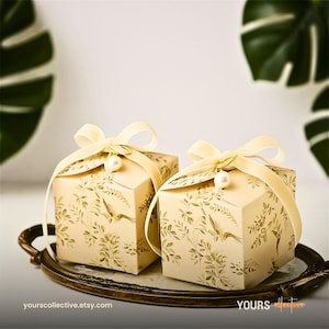 May include: Two cream-coloured gift boxes with a bird and floral pattern. Each box is tied with a light yellow ribbon and a pearl charm. The boxes sit on a decorative tray. The website address yours collective.etsy.com is visible.