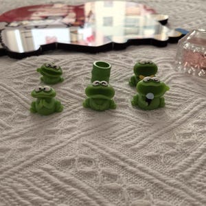 Könnte beinhalten: Eine Sammlung kleiner, grüner Froschfiguren mit weißen Augen. Ein Frosch hat ein Mikrofon, ein anderer eine Fliege und einer ist ein Rohr. Die Frösche sind auf einer weißen, strukturierten Oberfläche angeordnet.