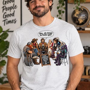 May include: A light gray t-shirt featuring a graphic of a diverse group of historical and futuristic figures sitting in a circle. A small chalkboard sign reads "TIME TRAVELERS SUPPORT GROUP" and a speech bubble says "Okay... who changed the timeline again?"