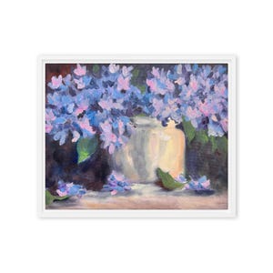 Puede incluir: Pintura enmarcada de un bodegón con un jarrón blanco lleno de hortensias azules y rosas. La obra de arte tiene un estilo suave y pictórico con pinceladas visibles y un fondo oscuro. Hojas verdes están esparcidas alrededor del jarrón.