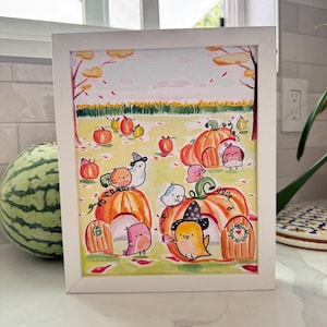 May include: A framed illustration in a white frame featuring a whimsical autumn scene with orange pumpkin houses, small colorful birds, and falling leaves. The artwork depicts a field with yellow flowers under a pale sky, with birds wearing hats.