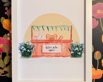 May include: A framed art print featuring a whimsical illustration of a cat behind a bake sale stand. The stand is painted in shades of orange and pink, with a banner of green triangles. The words "bake sale open" are written on the front.