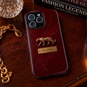 May include: A dark red, marble-patterned smartphone case featuring a gold-toned leopard figurine and a rectangular gold-toned metal plaque engraved with the name Seraphine, positioned centrally on the back of the device case.
