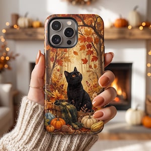 Puede incluir: Una funda para smartphone con una ilustración detallada de un gato negro sentado en una cesta de madera rodeado de follaje otoñal, calabazas y un pequeño ratón. El diseño incluye tonos cálidos de naranja, amarillo y marrón.