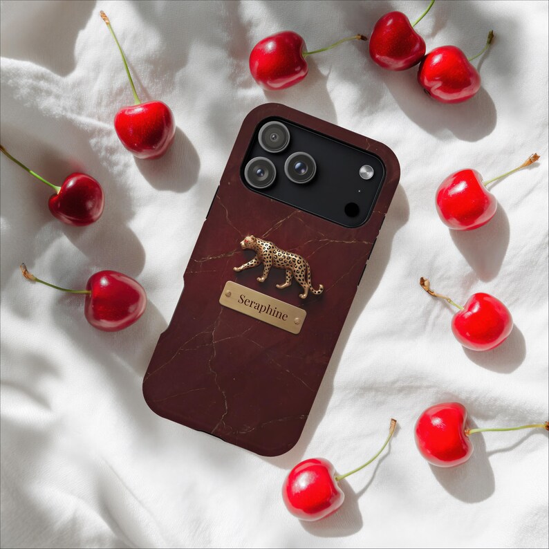 May include: A dark maroon smartphone case with a marble-like texture, featuring a gold-toned leopard figurine and a rectangular gold nameplate engraved with the text Seraphine, surrounded by several bright red cherries on a white fabric surface.