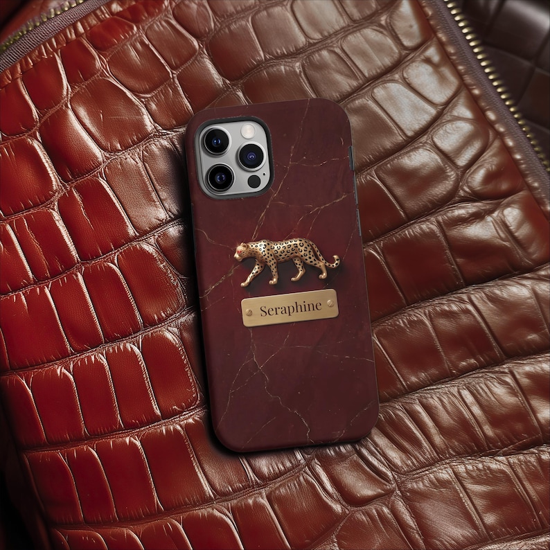 May include: A dark maroon phone case with a marble-like texture, featuring a gold-toned leopard figurine and a rectangular gold nameplate engraved with the text Seraphine. The case has a cutout for a triple-lens camera system on the top left.