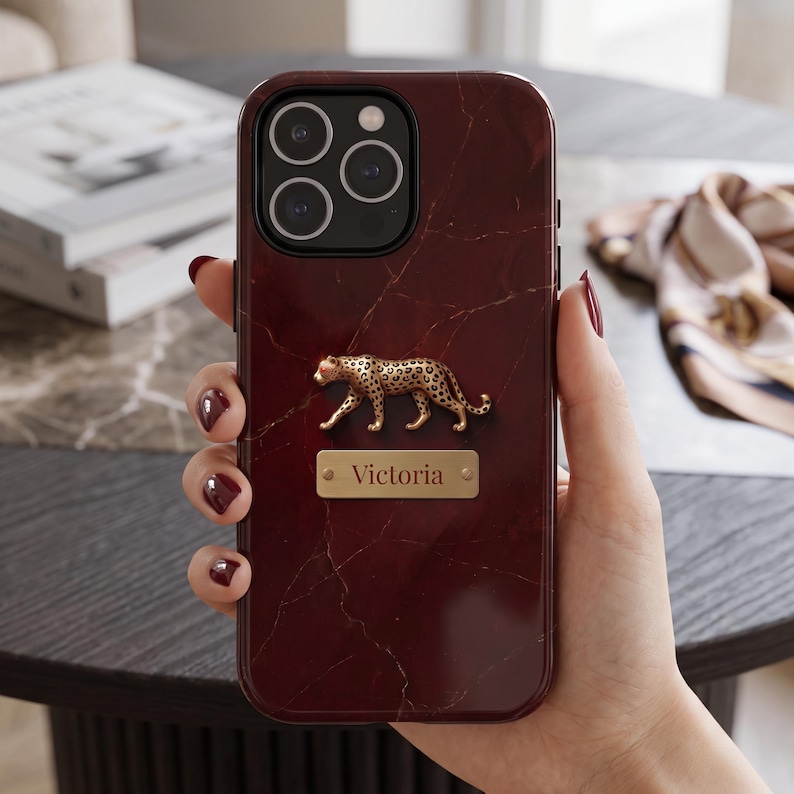 May include: A dark burgundy phone case with a white marble-like vein pattern. The case features a three-dimensional gold-toned leopard figure positioned above a rectangular gold-toned metal plaque engraved with the name Victoria in black text.