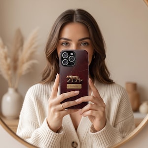 May include: A dark maroon phone case with a marble texture, featuring a gold-toned leopard figurine and a rectangular gold nameplate engraved with the name Isabella. The case includes a circular camera cutout with three lenses and a flash.