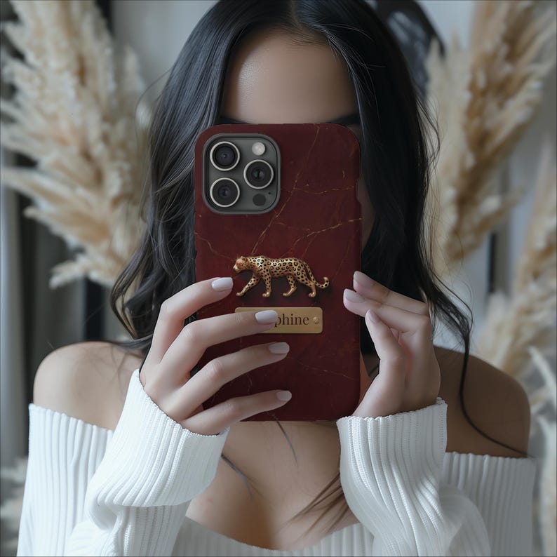 May include: A dark red phone case with a marble-like texture and thin gold veins. The case features a three-dimensional gold leopard figure positioned above a rectangular gold nameplate with the text "phine" engraved in a dark serif font.