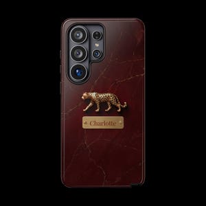 May include: A dark red smartphone case with a marble pattern, featuring a gold-toned leopard figurine and a rectangular gold-toned nameplate with the text Charlotte engraved in the center, positioned below the camera lens array on the back.