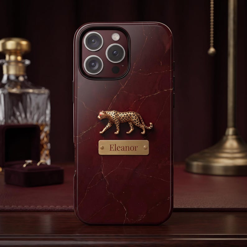 May include: A dark burgundy phone case with a marble texture and gold veining. The case features a three-dimensional gold leopard figure positioned above a rectangular gold nameplate engraved with the name Eleanor in a serif font.