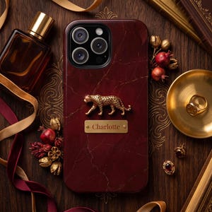 May include: A dark red marble-patterned phone case featuring a gold-toned leopard figurine and a rectangular gold nameplate with the text Charlotte engraved in the center. The case has a triple-lens camera cutout at the top and a smooth finish.