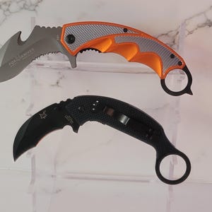 May include: Two karambit knives. The top knife has an orange and silver handle with a serrated blade and the text "FOX KNIVES". The bottom knife is all black.